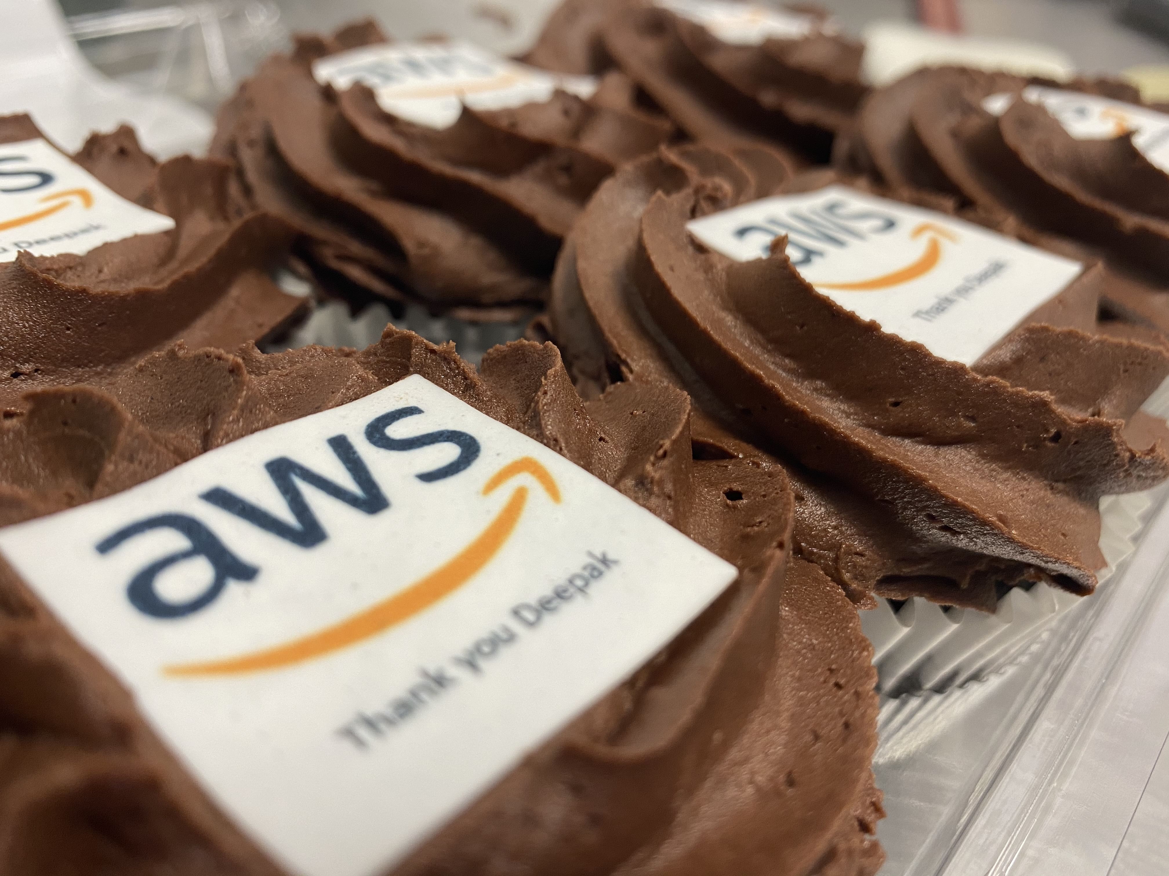 Close-up of chocolate cupcakes topped with custom-printed logos of 'AWS' and a thank you message. The edible masterpieces feature the text 'Thank you Deepak'. The personalised cupcakes are beautifully arranged in rows on a tray.